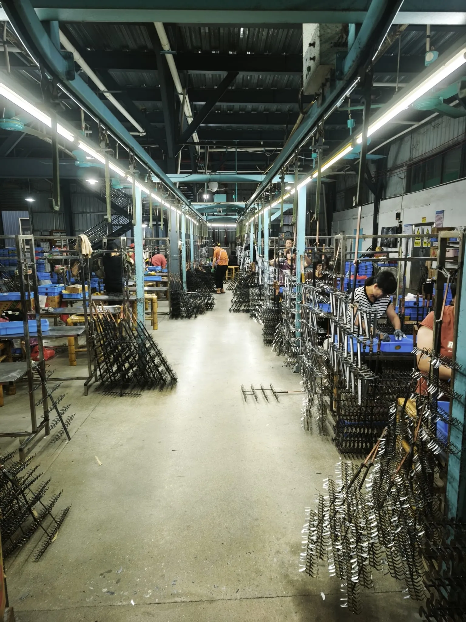 Operators at benches beside electroplating racks loaded with metal parts along a long factory aisle